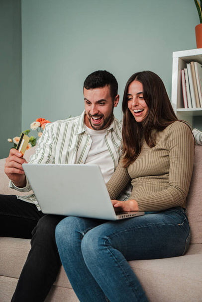 Vertical Feliz joven pareja caucásica adulto navegando por Internet utilizando un ordenador portátil para buscar una venta en casa. Emocionados marido y mujer sonriendo, buscando un nuevo apartamento sentado en un sofá en la sala de estar - Foto, imagen