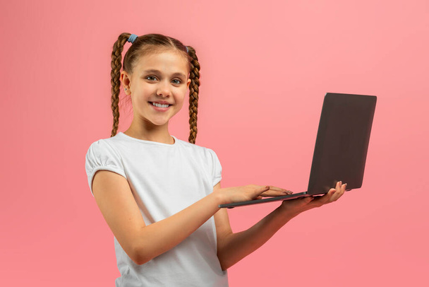 Een vrolijk jong meisje met gevlochten haar staande tegen een roze achtergrond trots met een open laptop - Foto, afbeelding