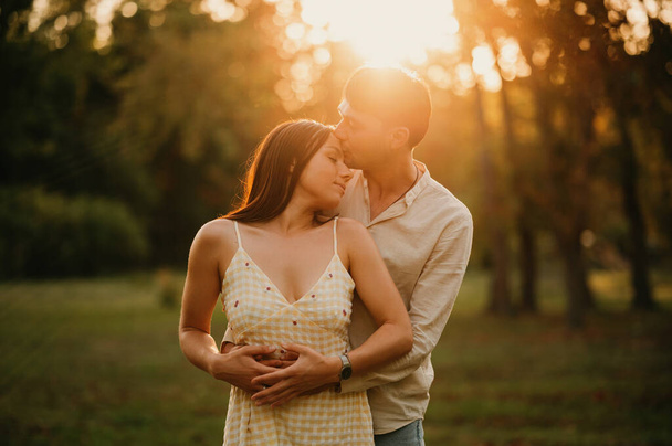 Νεαρός όμορφος άντρας φιλάει απαλά τη γυναίκα του στο μέτωπο ενώ την αγκαλιάζει το ηλιοβασίλεμα στο πάρκο. - Φωτογραφία, εικόνα