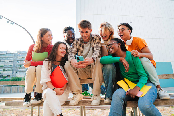 Skupina skutečných mladých lidí se baví spolu s mobilem, sedí na lavičce v univerzitním kampusu. Mnohonásobní přátelé sledující sociální média na chytrém telefonu. Teenage students using a phone - Fotografie, Obrázek