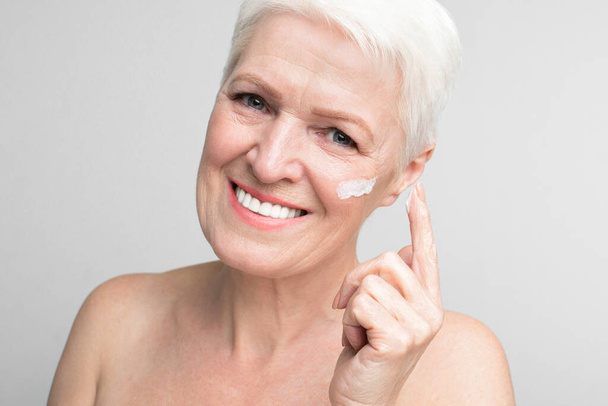 Весела літня європейська жінка, що застосовує крем для обличчя, відображає сутність s3niorlife та молодіжного духу - Фото, зображення