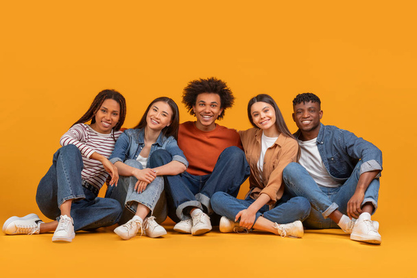 Eine zufriedene multiethnische Gruppe von Freunden, die lässig auf dem Boden zusammensitzen und ein engmaschiges internationales Team repräsentieren, isoliert auf orangefarbenem Hintergrund. - Foto, Bild