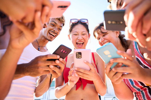Niski kąt widzenia grupy różnorodnych przyjaciół zebranych na plaży w kręgu z ich telefonów komórkowych. Pokolenie z osób korzystających z komórek wesoły i śmiech udostępniania śmieszne filmy w wakacje - Zdjęcie, obraz