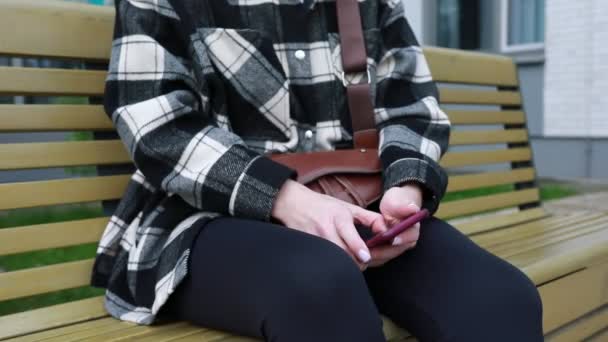 平らなジャケットと黒いズボンを着た若い女性は,木製の公園のベンチに座っている. 彼女の焦点は,彼女の指が画面をすばやく移動するので,彼女のスマートフォンにあります, おそらく作曲 - 映像、動画
