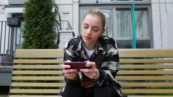 Μια στοχευμένη νεαρή γυναίκα, που κάθεται σε ένα ηλιόλουστο παγκάκι του πάρκου, ασχολείται με το smartphone της, περιδιαβαίνοντας το περιεχόμενο με μια ματιά συγκέντρωσης. Η περιστασιακή ενδυμασία της προτείνει μια χαλαρή έξοδο καθώς περνάει χρόνο. - Πλάνα, βίντεο