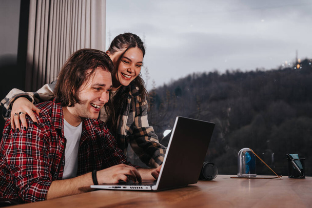 Sonriendo joven hombre y mujer usando un ordenador portátil juntos en casa, compartiendo un momento de alegría. - Foto, imagen