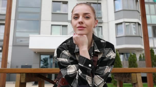 A woman is seen sitting on a bench in front of a building. She appears relaxed and is observing her surroundings. The building behind her is tall and modern, adding to the urban setting of the scene. - Footage, Video