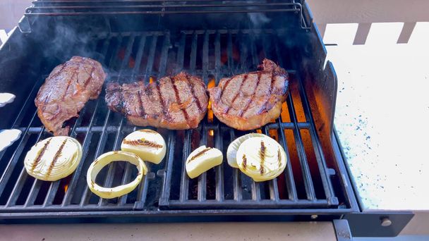 Ribeye steaks are sizzling alongside golden grilled onions on a barbecue grill, with wisps of smoke hinting at the flavorful feast being prepared.  - Photo, Image