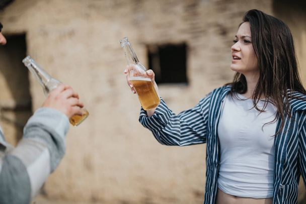 Eine junge Frau und ein Mann, die Bierflaschen in der Hand halten und im Freien stehen, wobei die Frau während eines lockeren Urlaubsmoments unsicher oder unzufrieden wirkt. - Foto, Bild