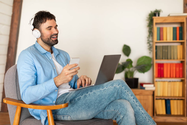 Un uomo allegro è seduto comodamente su una poltrona, indossando un paio di cuffie over-the-ear mentre si impegna con il suo computer portatile e tenendo uno smartphone bianco - Foto, immagini