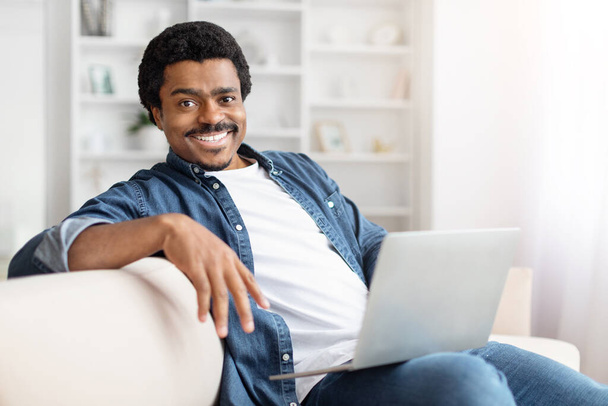 En las comodidades del hogar, un hombre afroamericano disfruta de un tiempo libre con su computadora portátil, luciendo una sonrisa de bienvenida - Foto, Imagen