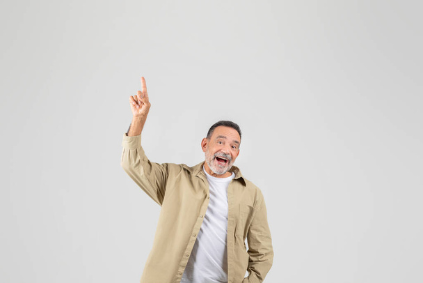 Un hombre mayor de pie con una chaqueta bronceada y una camisa blanca con una expresión neutra en su cara, señalando con el dedo hacia arriba en el espacio de copia - Foto, imagen