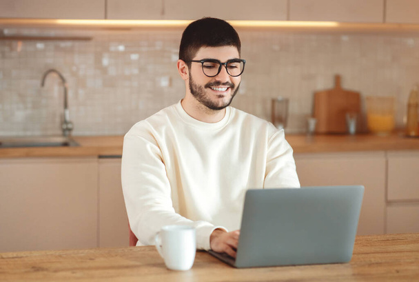 Een man zit aan een tafel, gefocust op het gebruik van zijn laptop computer. Hij blijkt intensief te werken of te studeren, typt en klikt op het laptopscherm. De achtergrond is eenvoudig en onbeschrijflijk. - Foto, afbeelding