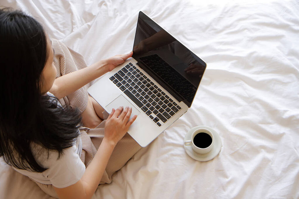 top view of Asian Woman sitting on the bed with cup of coffee and working on her laptop. with copy space - Photo, Image