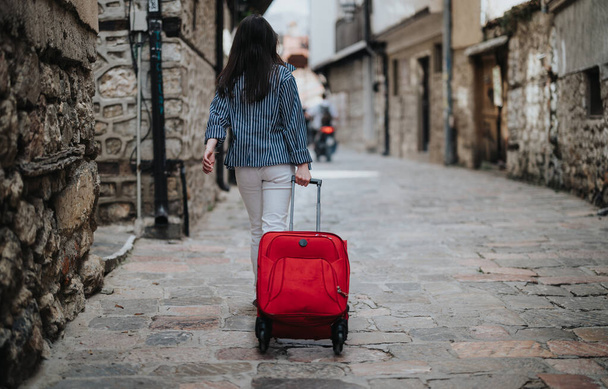 Vista trasera de una joven turista paseando con equipaje por un pintoresco callejón de la ciudad en un día soleado - Foto, Imagen