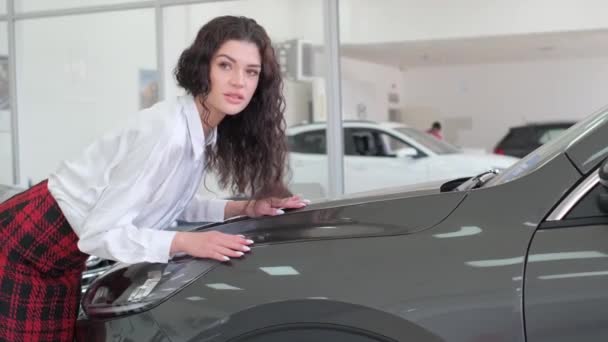 Привлекательная темноволосая женщина позирует перед камерой возле нового автомобиля, опираясь на капот автомобиля. Видеосъемка брюнетки с красивыми волосами - Кадры, видео
