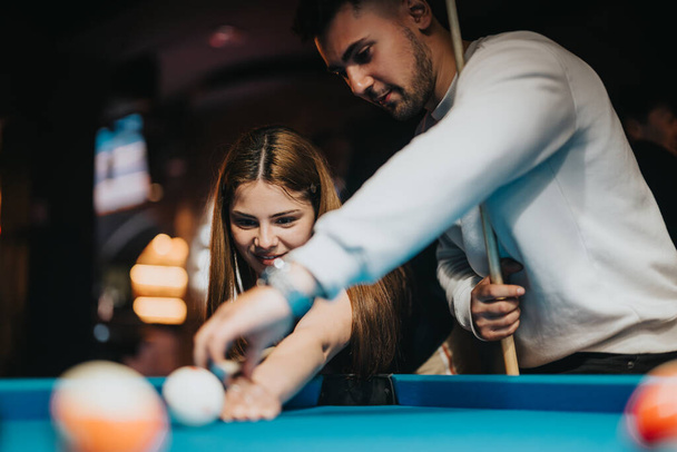 A man and woman share a playful moment while playing pool, surrounded by the warm ambiance of a billiard hall. - Photo, Image
