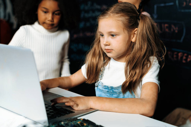 Beyaz önlüklü sarışın kız BTMM sınıfında kendine güvenen dizüstü bilgisayar kullanarak robot teknolojisini öğreniyor. Beyaz süveter giyen kız meraklı gözlerle kodlamasına bakıyor. Etkinlik. - Fotoğraf, Görsel
