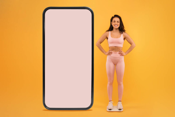 A cheerful woman dressed in pink fitness clothes confidently stands on scales beside an oversized smartphone with a blank screen, slimming tracker - Photo, Image