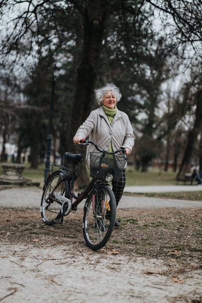 Una mujer mayor activa con una sonrisa brillante se para con su bicicleta en un parque tranquilo, encarnando un estilo de vida saludable. - Foto, imagen