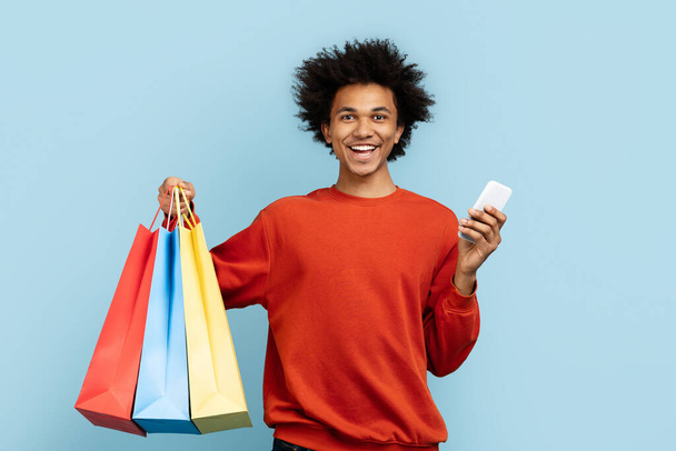 Esta imagen muestra a un tipo de zoom negro radiante de felicidad mientras sostiene bolsas de compras y un teléfono inteligente, aislado sobre un telón de fondo azul - Foto, imagen