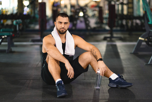 Muž v atletických šatech sedící na podlaze tělocvičny, obklopený cvičební výstrojí. Zdá se, že si dává pauzu nebo se po tréninku protáhne.. - Fotografie, Obrázek