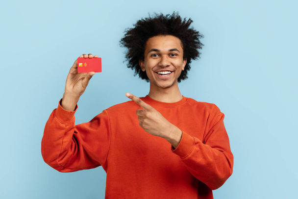 Egy afro-amerikai fickó boldogan mutat a piros hitelkártyájára, kék háttérrel elszigetelve, ideális pénzügyi koncepciókhoz. - Fotó, kép