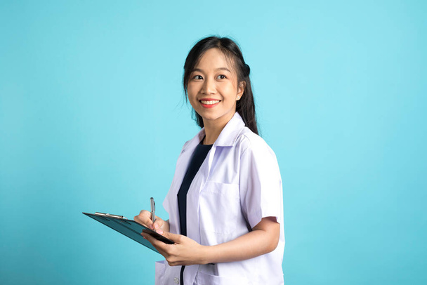Hymyilevä nuori nainen Aisan lääkäri tai sairaanhoitaja, joka tekee muistiinpanoja leikepöydällä seisoen sinistä taustaa vasten  - Valokuva, kuva