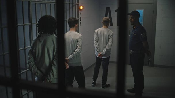 Un grupo de prisioneros está cerca de la celda de la cárcel, el alcaide habla con ellos. Adolescentes multiétnicos cumplen condena de prisión en un centro de detención. Jóvenes criminales en prisión. Sistema de justicia. Vista a través de barras de metal. - Foto, imagen