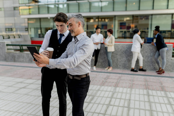 Senior zakenman deelt inzichten met een jonge mannelijke collega via een digitale tablet, tijdens een informele bijeenkomst buiten. - Foto, afbeelding