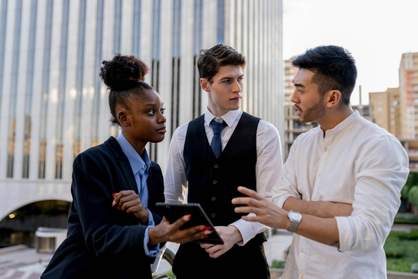 Trois jeunes professionnels diversifiés engagés dans une discussion sérieuse, utilisant une tablette numérique pour relever des défis complexes dans un quartier d'affaires de la ville. - Photo, image