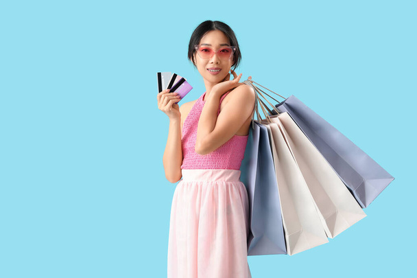Glücklich schöne junge Asiatin mit Einkaufstaschen und Kreditkarten auf blauem Hintergrund - Foto, Bild