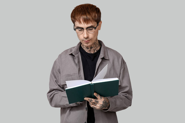 Молодой татуированный человек читает книгу на сером фоне - Фото, изображение