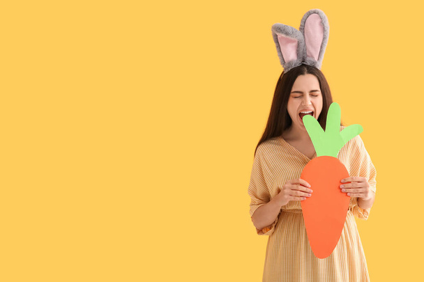 Krásná mladá šťastná žena v velikonoční zajíček uši s papírovou mrkví na žlutém pozadí - Fotografie, Obrázek