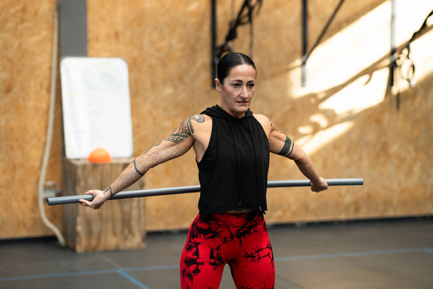 Horizontales Foto mit Kopierraum einer starken tätowierten Frau, die sich in einem Fitnessstudio mit einer Stange dehnt - Foto, Bild