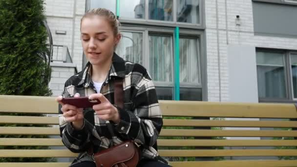 Молода жінка захоплена своїм смартфоном, сидячи на дерев'яній лавці поза сучасною будівлею. Здається, це приємний весняний день, і вона, здається, є або смс, перегляд, або - Кадри, відео