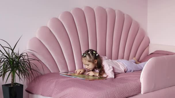 Uma menina está deitada na cama olhando para o livro. O que escolher um livro ou um smartphone. O conceito de infância e lazer. Filmagem 4k. - Filmagem, Vídeo