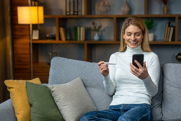Smiling and excited adult woman checking her recent pregnancy test, sitting on gray couch at home, copy space - Foto, Imagen