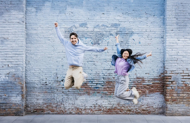 Junge Männer und Frauen springen an einer blauen Backsteinmauer vorbei. Paar springt fröhlich gegen eine farbige Wand und lächelt. - Foto, Bild