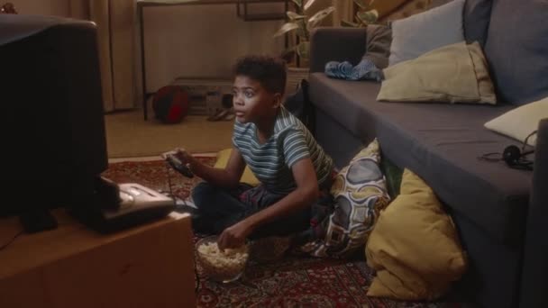 Side volledige handheld schot van Afro-Amerikaanse kind opgenomen spelen console arcade video game op vintage tv zitten op de vloer en verslindende popcorn alleen thuis - Video