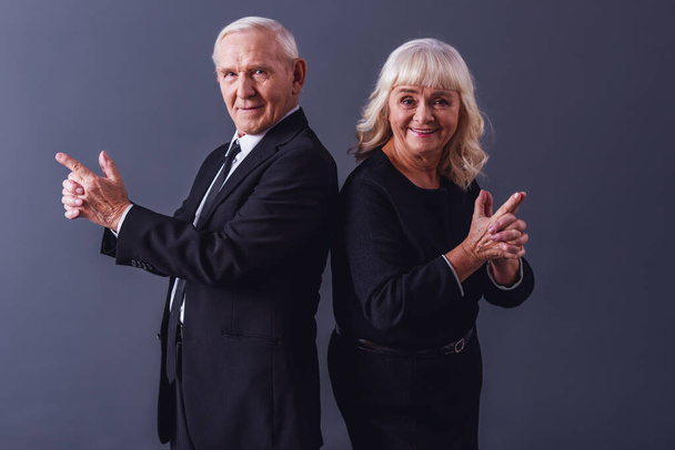 Krásný starý pár v elegantní oblečení je drží za ruce společně napodobující zbraně, při pohledu na fotoaparát a usmívá se na šedém pozadí - Fotografie, Obrázek