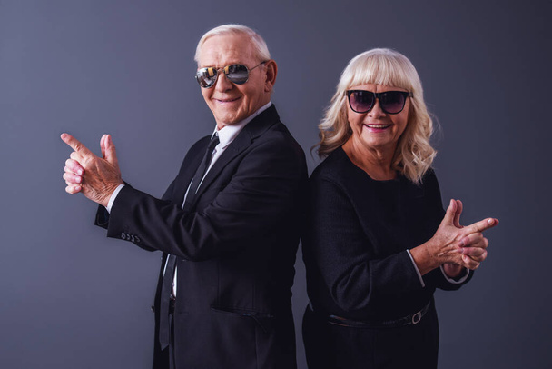 Beau vieux couple dans des vêtements élégants et des lunettes se tient la main ensemble imitant des fusils, regardant la caméra et souriant, sur fond gris
 - Photo, image
