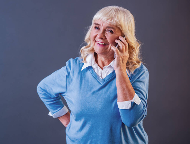 Gyönyörű régi hölgy alkalmi ruhák beszél a mobiltelefonján, és mosolyogva, szürke háttér - Fotó, kép
