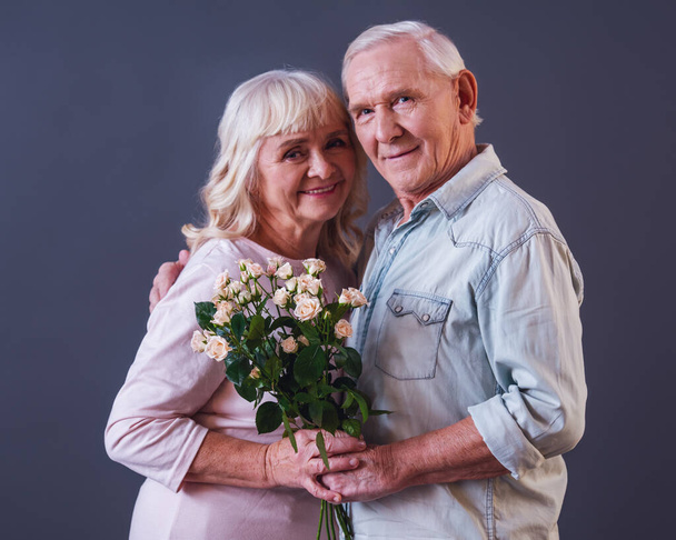 Mooie oude man en vrouw in casual kleding zijn houden van bloemen, kijken naar camera en glimlachend, op grijze achtergrond - Foto, afbeelding