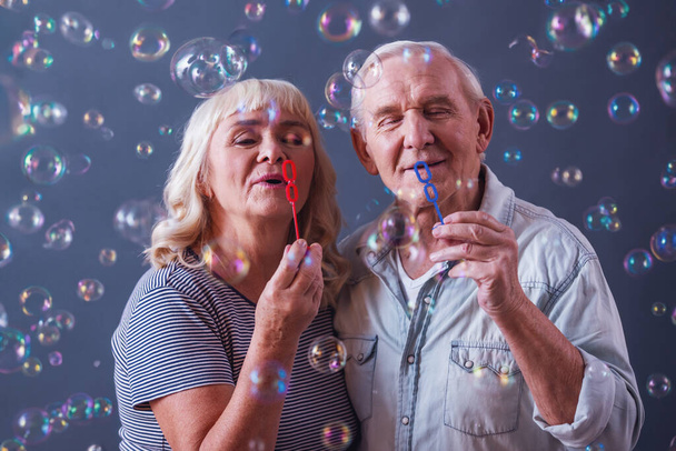 Mooie oude man en vrouw in casual kleding zijn blazen van zeepbellen en glimlachend, op grijze achtergrond - Foto, afbeelding