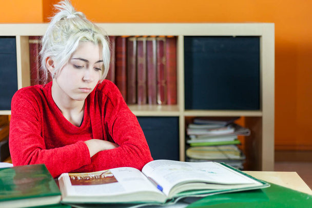 Egy vörös ruhás fiatal nő keményen tanul a könyvtári asztalnál, kimerülten néz ki könyvekkel és polcokkal a háttérben, kimerültséget mutat.. - Fotó, kép