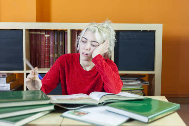 Mujer joven de cabello rubio, cansada, sosteniendo un bolígrafo mientras lee un libro en un entorno de biblioteca, rodeada de libros y mostrando signos de fatiga. - Foto, Imagen