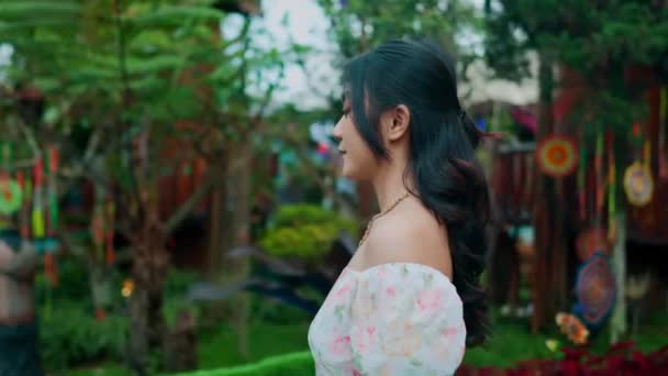Seitenansicht einer jungen Frau in einem floralen Kleid, die morgens einen üppigen Garten mit hängenden Dekorationen genießt - Filmmaterial, Video