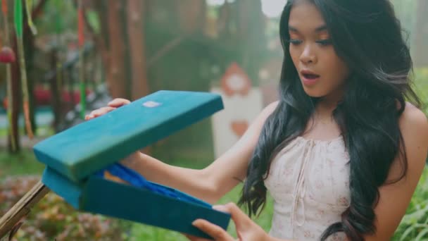 Mujer sonriente abriendo una caja de regalo con una tela azul en el interior, al aire libre con vegetación en el fondo durante el día - Metraje, vídeo