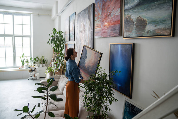 Interested female artist holds painting in hands and searches for suitable place on wall of home to hang result of creative work. Thoughtful designer woman decoration art studio with oil picture.  - Photo, Image